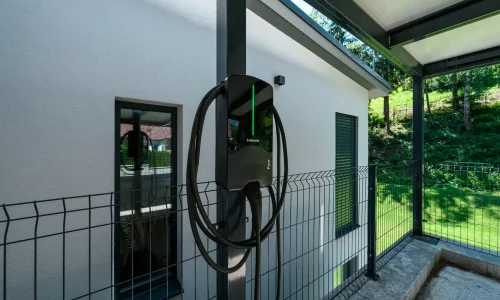 Elektroauto-Ladestation am Pfosten des anthrazitfarbenen Carports an verputzter Fassade.