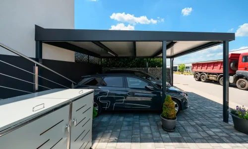 Dunkler Flachdach-Carport schützt Fahrzeuge an gepflasterter Einfahrt.