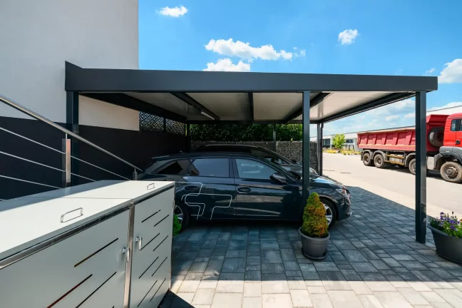 Dunkler Flachdach-Carport schützt Fahrzeuge an gepflasterter Einfahrt.