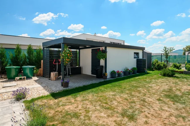 Carport mit anthrazitfarbener Metall-Lamellen-Verkleidung an weiß verputztem Gartenhaus.