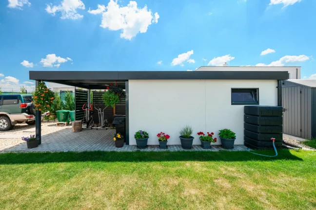 Offener Carport mit weiß verputztem Gartenhaus und anthrazitfarbenem Flachdach im Garten.