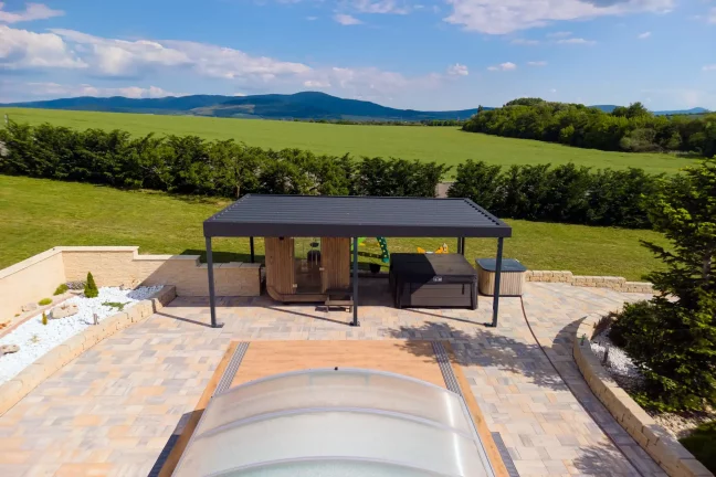 Anthrazitfarbene Pergola mit Gartenmöbeln neben einem Pool mit transparenter Abdeckung, grüne Hügel im Hintergrund