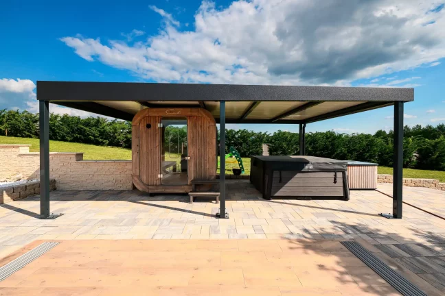 Anthrazitfarbene Pergola mit Flachdach neben einer Fasssauna auf einer gepflasterten Terrasse, blauer Himmel