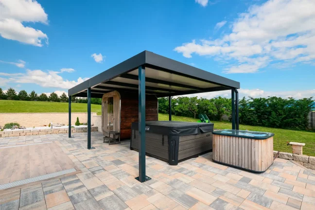 Anthrazitfarbene Pergola mit Whirlpool darunter auf einer gepflasterten Terrasse, grüner Garten und blauer Himmel