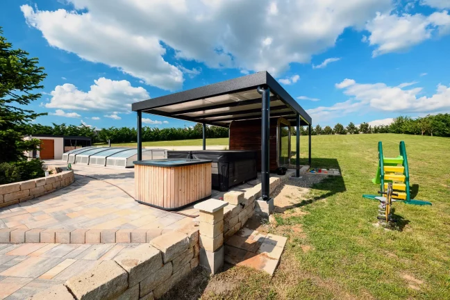 Anthrazitfarbene Pergola mit Whirlpool und Kinderspielplatz im Hintergrund, gepflasterte Terrasse am Garten