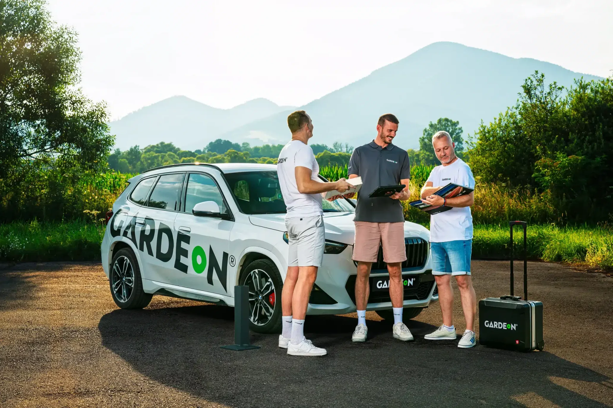 Drei GARDEON-Agenten besprechen Unterlagen am Firmen-BMW in der Natur.