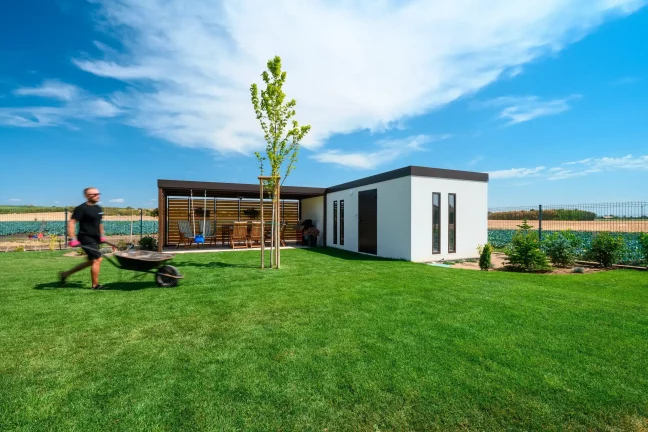 Mann mit Schubkarre geht durch den Garten am Gartenhaus mit Überdachung vorbei