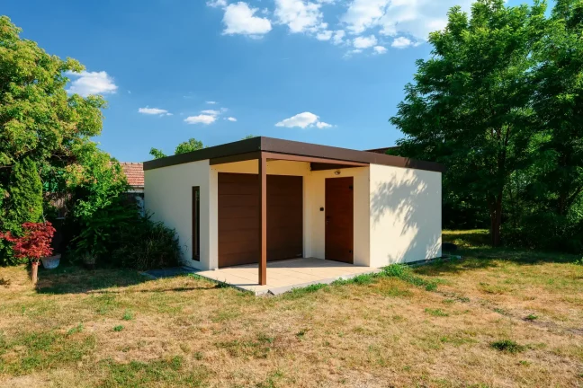 Gartenhaus mit braunem Garagentor und überdachter Terrasse zwischen Bäumen