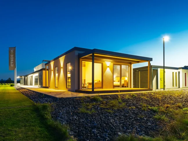 Vordere Ecke des beleuchteten Tiny Houses mit überdachter Terrasse in der Dämmerung