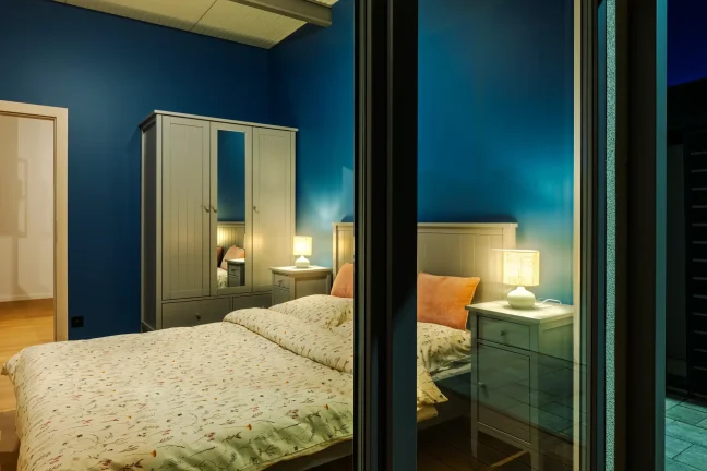 Schlafzimmer des Tiny Houses mit eingeschalteten Lampen, Blick durchs Fenster in der Dämmerung